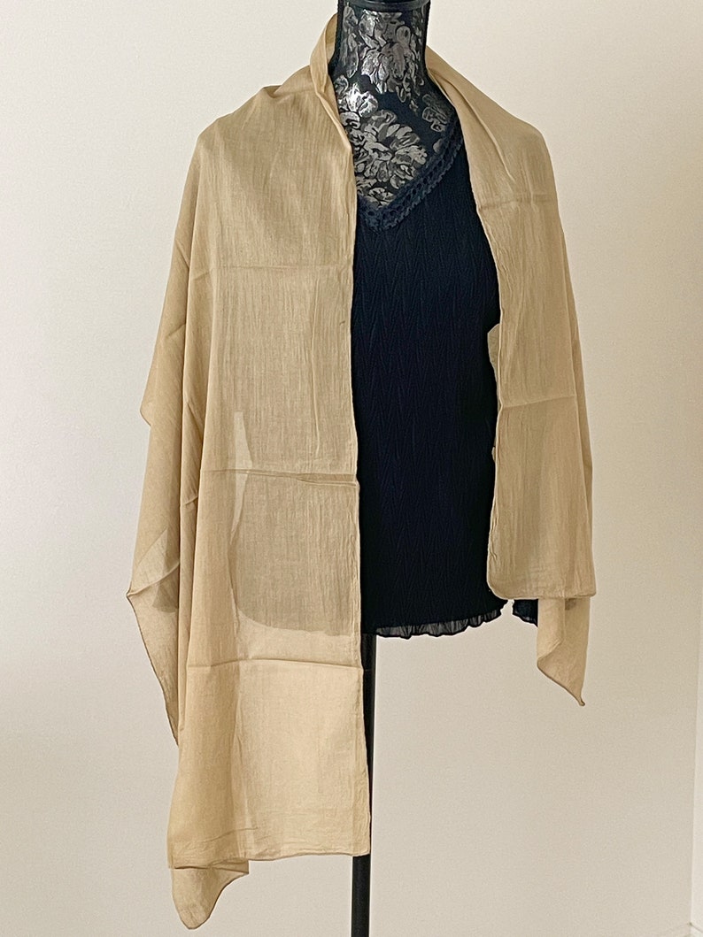 May include: A light beige scarf made of a soft, sheer fabric. The scarf is draped over a mannequin and is folded in half.
