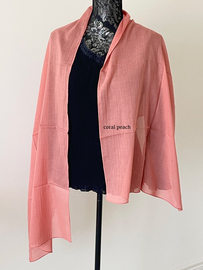 May include: A coral peach colored scarf made of a lightweight fabric. The scarf is draped over a black mannequin.