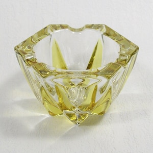 May include: A clear glass ashtray with a yellow tint and a geometric design. The ashtray has a hexagonal shape and a wide, shallow bowl.