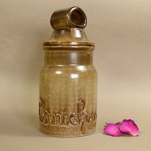 May include: A brown ceramic jar with a lid and a handle. The jar has the word "Kornichorn" written on it in brown lettering.