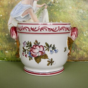 May include: White ceramic planter with floral designs in pink, green, and blue. The planter has two handles shaped like stylized flowers. The planter is sitting on a green surface.