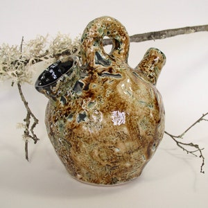 May include: A handcrafted ceramic pitcher with a unique design. The pitcher features a textured surface with brown, green, and cream tones. It has a spout, a handle with a hole, and a second smaller spout. Decorative twigs and lichen are in the background.