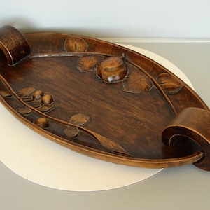 May include: Oval wooden serving tray with carved leaf and fruit details. The tray has a dark brown finish and curved handles at each end. The tray is placed on a white and gray surface.