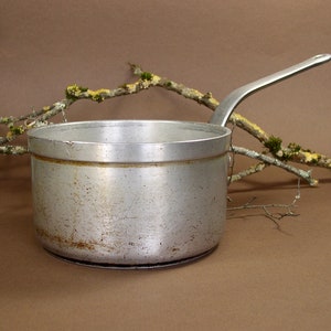May include: A small, well-used aluminum saucepan with a long, curved handle. The pot shows signs of wear and tear, with some discoloration. A branch with lichen and moss is in the background, adding a natural element to the composition.
