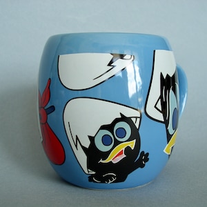 Peut inclure: Une tasse en céramique bleue avec un motif de dessin animé représentant un personnage noir et blanc avec trois yeux et une moustache jaune. Le personnage est entouré d'autres personnages de dessin animé, dont un oiseau blanc avec un bec noir et un personnage rouge et blanc avec un chapeau rouge.