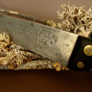 Peut inclure: Gros plan sur un couteau vintage avec une lame argentée usée et un manche noir. La lame porte du texte et un logo, et est ornée de trois étoiles. Le manche est fixé avec des rivets en laiton. Le couteau repose sur une petite branche avec du lichen séché.