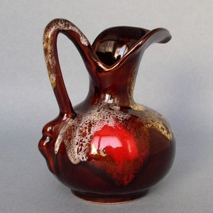 Op de afbeelding: Een bruine keramische kan met een rode en gouden glazuur. De kan heeft een brede opening en een gebogen handvat.