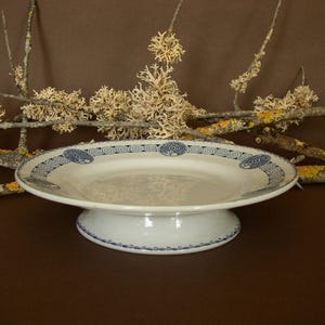 May include: A white ceramic cake stand with a blue and white patterned rim. The stand has a pedestal base and is perfect for serving desserts or snacks.