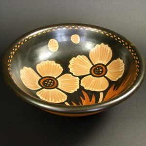 May include: A black ceramic bowl with a brown rim and two stylised flowers painted in beige on the inside. The flowers have brown centres and are surrounded by brown leaves.