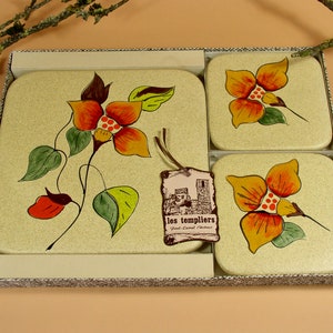 May include: Set of four beige ceramic coasters with a floral design. Each coaster features a yellow, orange, and red flower with green leaves. The coasters are packaged in a beige box with a brown tag that reads "les templiers" and "Hotel-Cafe-Restaurant".
