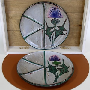 May include: Two ceramic plates with a divided section design. Each plate features a green and purple thistle flower design.
