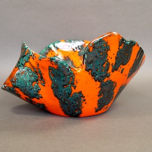 May include: A handmade ceramic bowl with an orange base and dark teal speckled pattern. The bowl has a unique, folded shape with raised edges. The interior is a glossy orange, and the exterior has a textured, speckled finish.