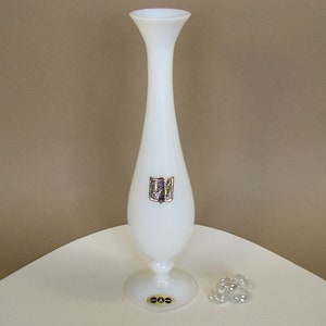May include: A white ceramic vase with a gold emblem on the front. The vase has a slender, elongated shape and a round base. It is sitting on a white surface.