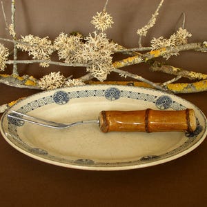 Peut inclure: Un plat de service ovale avec une bordure décorative bleue et blanche, contenant une fourchette de service avec une poignée en forme de bambou. Le plat est de couleur crème. Des brindilles avec du lichen sont en arrière-plan.
