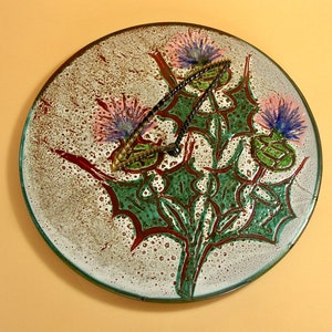 May include: A round ceramic plate with a white background and a green, red, and brown design of three stylised thistles with purple and blue flowers.