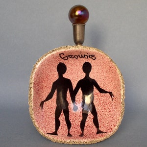 Op de afbeelding: Een roze keramische schaal met een gouden rand en een zwart silhouet van het sterrenbeeld Tweelingen. De schaal heeft een kleine, iriserende glazen knop bovenop. Het woord "Tweelingen" staat in het zwart op de schaal.