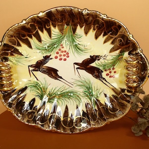 May include: A large, oval ceramic serving dish with a cream-colored background and a gold rim. The dish is decorated with a hand-painted design of two deer in a forest setting with pine branches and red berries.