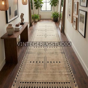 May include: Beige and black patterned runner rug with a geometric design. The rug features a central vertical pattern with horizontal lines and diamond shapes, and a fringed edge. The rug is displayed in a hallway with wooden floors and walls.