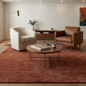 May include: A modern living room featuring a large, rectangular rust-colored rug. Two armchairs, one cream-colored and one brown leather, are arranged around a round wooden coffee table. The room is decorated with plants and abstract art.