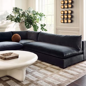Peut inclure: Un canapé d'angle gris foncé avec un coussin blanc et un coussin rond brun. Le canapé est dans un salon avec une table basse blanche et un tapis à motifs. Il y a une grande plante en pot en arrière-plan.