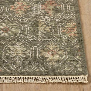 Puede incluir: Primer plano de una sección de una alfombra verde con un patrón geométrico detallado en tonos verdes, beige y coral. La alfombra tiene una franja a lo largo del borde inferior, agregando un elemento decorativo al diseño.