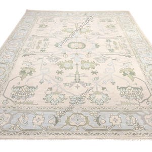 Puede incluir: Una alfombra rectangular grande con un intrincado patrón floral y geométrico. La alfombra presenta un fondo color crema con acentos de azul claro y verde. El borde tiene un diseño repetitivo.