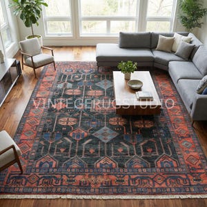 May include: A large, rectangular area rug with a dark teal field and a border in shades of orange and blue. The rug features geometric patterns and is placed in a living room with a gray sectional sofa and wooden coffee table.