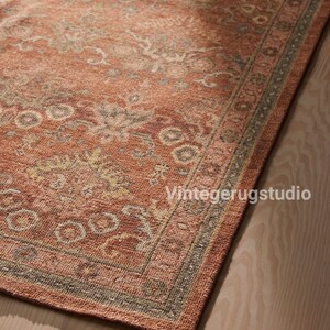 May include: A corner view of a distressed, rectangular rug with a faded, floral pattern in shades of rust, beige, and olive green. The rug has a border with a repeating geometric design. The text "Vintegerugstudio" is visible in the bottom right corner.