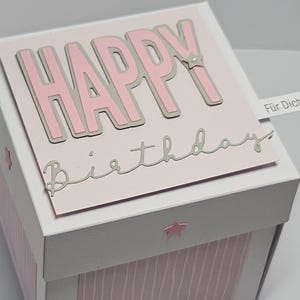 Könnte beinhalten: Eine weiße und rosa Geburtstagsgeschenkbox mit den Worten "HAPPY Birthday" in rosa und silbernen Buchstaben. Die Box hat ein gestreiftes Muster und Sternakzente. Ein kleines Banner lautet "Für Dich".