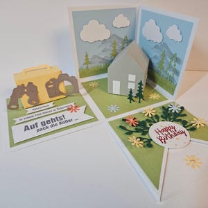 Könnte beinhalten: Ein mehrteiliges Pop-up-Grußkartenset mit einem gelben Koffer, Fernglas- und Kamera-Ausschnitten, einem grauen Haus in einer Berglandschaft sowie einer floralen Geburtstagskarte mit dem Text "Happy Birthday" und "Auf gehts! pack die Koffer".