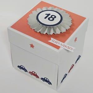 Könnte beinhalten: Eine weiße, quadratische Geschenkbox mit einem korallenfarbenen Deckel, einer grauen Papierrosette und der Zahl 18 in der Mitte. Die Seiten sind mit kleinen korallenfarbenen Sternen, einem Banner mit dem Text GLÜCKWUNSCH und kleinen blauen und roten Autos verziert.