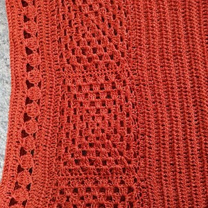 Może przedstawiać: Zbliżenie na tkaninę wykonaną na szydełku z fakturowanym wzorem. Tkanina jest ciemno pomarańczowa i ma powtarzający się wz&oacute;r kwadrat&oacute;w i k&oacute;ł.