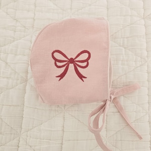May include: A pale pink baby bonnet featuring a burgundy bow embroidery. The bonnet, with a tie closure, is crafted from a soft, textured fabric. It rests on a white, quilted surface, offering a delicate aesthetic.