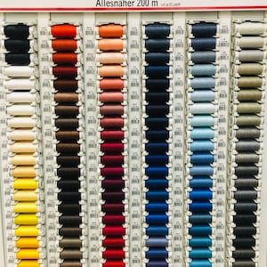 Könnte beinhalten: Eine Wandanzeige mit Gutermann Allesnäher 200 m Garnrollen in verschiedenen Farben, darunter Schwarz, Rot, Blau, Braun, Gelb und Grau. Die Rollen sind in Reihen und Spalten angeordnet, wobei jede Rolle mit einer Nummer gekennzeichnet ist.