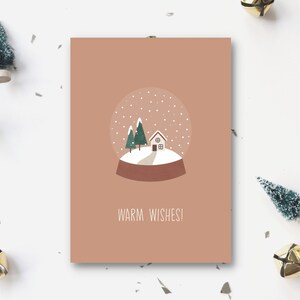 以下が含まれることがあります： 雪だるまの絵が描かれた茶色のカード。雪だるまは小さな家と2本の木があり、雪が降っている。雪だるまの下には「Warm Wishes!」という文字が書かれている。