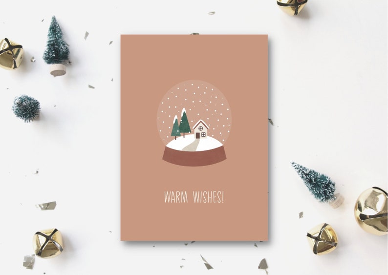 以下が含まれることがあります： 雪だるまの絵が描かれた茶色のカード。雪だるまは小さな家と2本の木があり、雪が降っている。雪だるまの下には「Warm Wishes!」という文字が書かれている。