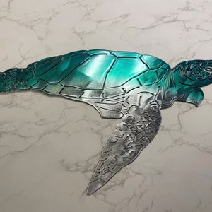 May include: A teal and silver metal sea turtle wall decor. The turtle is in a swimming position with its head turned to the right. The turtle's shell is teal and the rest of the turtle is silver.
