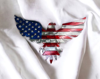 May include: A metal eagle sculpture with the American flag design. The eagle is in a spread-wing pose with its head facing forward. The sculpture is made of metal and has a distressed finish.