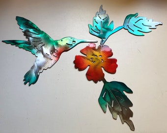 May include: Metal hummingbird sculpture with a gradient of colours, including green, blue, red, and silver. The hummingbird is depicted in flight, approaching a flower with similar colour gradients. Leaves are also included in the design.