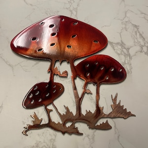 May include: A metal wall art piece featuring three stylized mushrooms in shades of red and copper. The mushroom caps have small, evenly spaced holes. The artwork is mounted on a white marble-patterned surface.