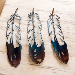 May include: Three metal feathers with a blue and brown ombre finish. The feathers are cut out in a detailed design and have a rustic look.