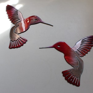 Może przedstawiać: Dwa metalowe kolibry z czerwono-srebrnym gradientem. Kolibry są w locie z rozłożonymi skrzydłami.