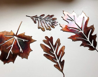 May include: Four metal leaf cutouts in various shapes and sizes. The leaves are a copper color with a reddish-brown patina. The leaves are arranged on a white surface.
