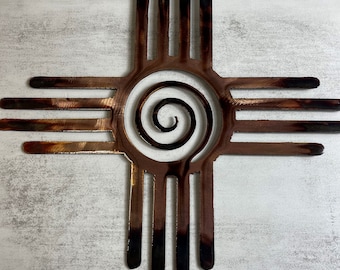 May include: A copper-colored metal wall art piece depicting the traditional Native American symbol of the "Sun" or "Four Directions". The symbol is a stylized cross with four arms, each with four lines, and a spiral in the center.