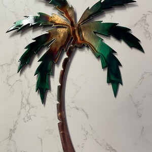 May include: A metal palm tree wall decor with a green and brown finish. The palm tree has a textured, rustic look.