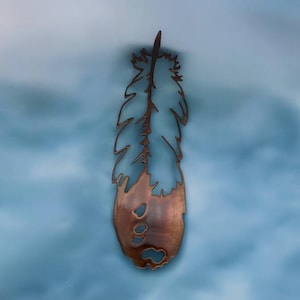May include: A copper-colored metal feather cutout with a rustic finish. The feather is a decorative wall hanging.