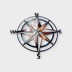 May include: A metal compass rose with a star design, featuring the cardinal directions: North, South, East, and West. The compass has a dark, metallic finish with a circular frame and a starburst center. The design is suitable for wall decor.