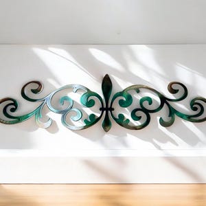 May include: A decorative metal wall art piece with a green and brown finish. The design features a stylized fleur-de-lis in the center with scrolling floral patterns on either side.