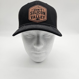 May include: Black trucker hat with a mesh back and a curved brim. The front features a brown leather patch with the words "DON'T SILICON MY VALLEY" in white lettering, with a mountain and cabin graphic.