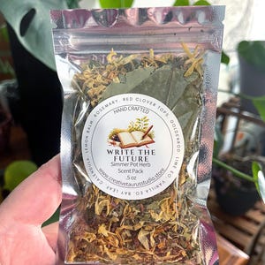 May include: A clear, sealed bag filled with dried herbs and botanicals. The bag features a circular label with the text "WRITE THE FUTURE Simmer Pot Herb Scent Pack." Ingredients include rosemary, red clover tops, and calendula. The bag is held by a hand.